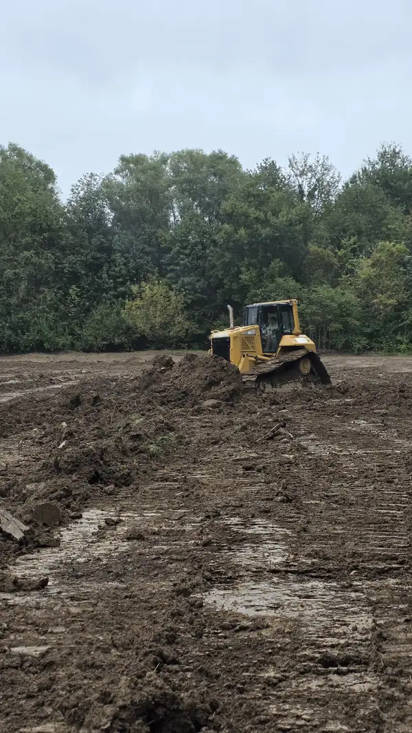 Bulldozer pushing a pile of dirt across a muddy job site with trees behind