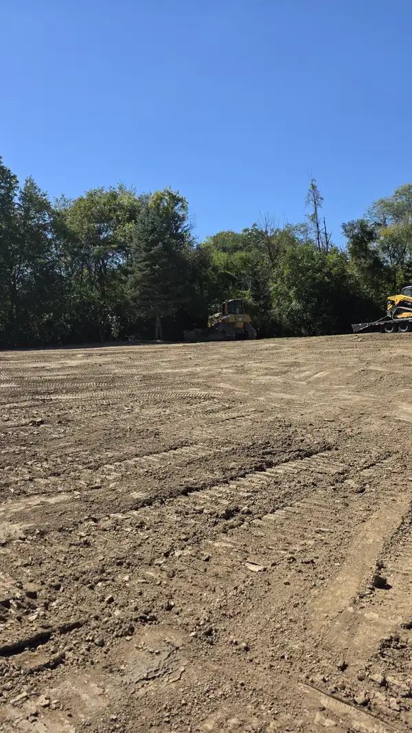 Freshly graded dirt lot with dozer track marks and equipment parked along the tree line