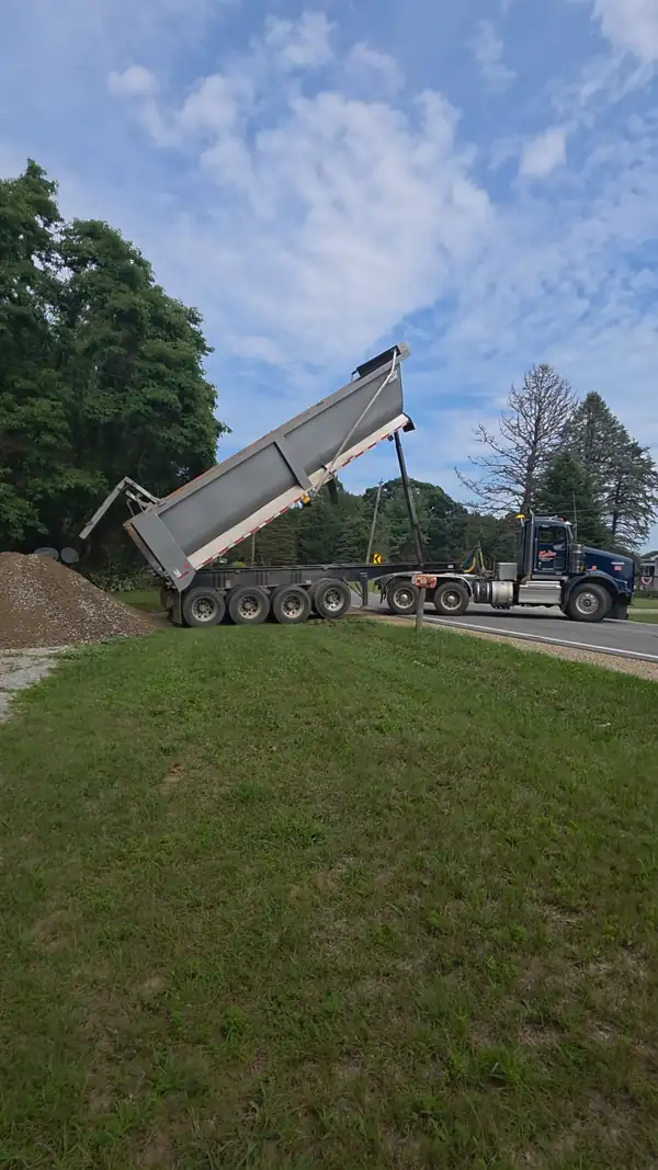 Dump trailer raised and unloading a pile of gravel at the edge of a property