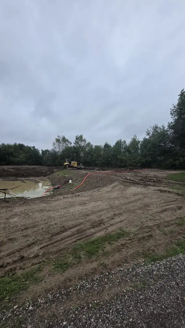 Pond excavation with standing water, a pump hose, and a bulldozer working the far bank